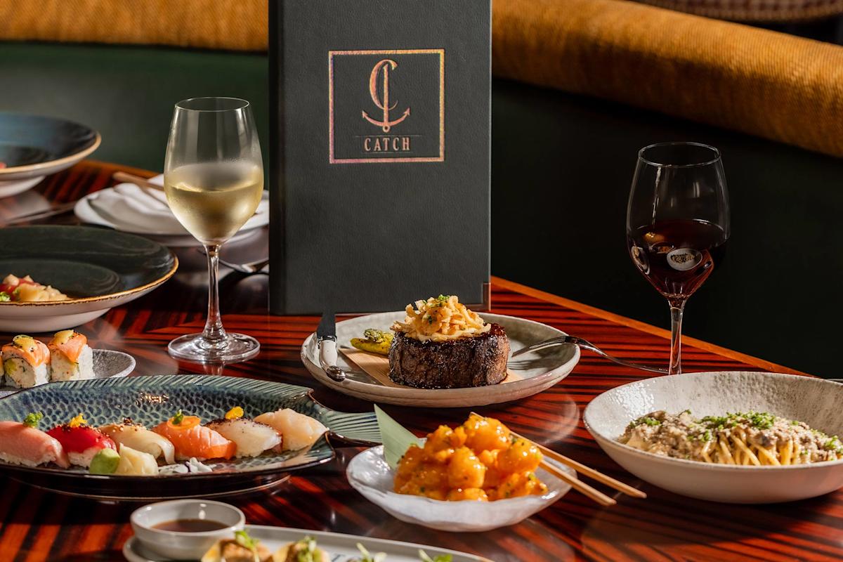 A table set with gourmet dishes including sushi, steak, pasta, and shrimp, accompanied by glasses of white and red wine, with a dark Catch menu upright in the center.