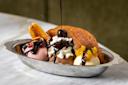 A banana split served in a silver dish with three scoops of ice cream topped with chocolate sauce, whipped cream, a cherry, and fruit garnishes. A waffle slice is placed on the side.