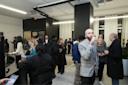 A group of people socialize at an indoor event in a modern office space; some are talking, others are drinking, with coats visible, suggesting a colder season. Bright lighting and contemporary decor are present.