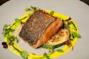 A piece of seared salmon with crispy skin sits on a slice of grilled zucchini, garnished with microgreens and herbs, and surrounded by a swirl of yellow sauce on a grey plate.