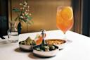 A table with a glass of iced tea garnished with orange slices, a small vase holding flowers, and three small bowls containing Romanesco broccoli, carrot sticks, mixed olives, and snacks.