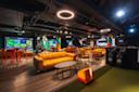 Modern indoor golf lounge with yellow sofas, tall red tables and stools, multiple golf simulators, golf bags, and colorful lighting, creating a relaxed and vibrant atmosphere.