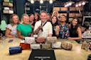 A group of people smiling and posing together behind a table displaying various handbags. A man in the center holds a drink and points toward the camera. The setting appears celebratory, with a backdrop of shelves containing bags and boxes.