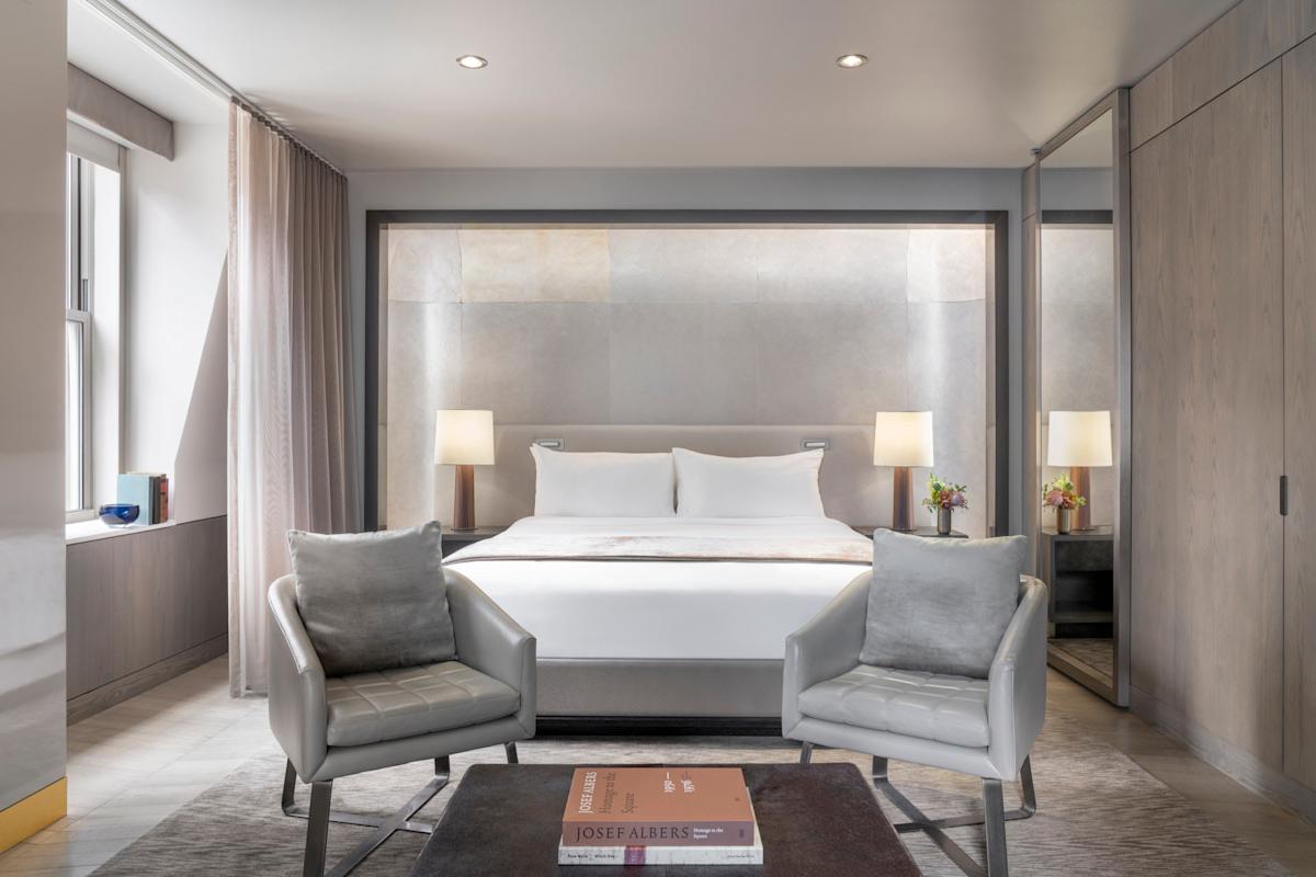 A modern hotel room with a large bed, two bedside tables with lamps, two gray armchairs, a small table with books, and a large mirrored wardrobe, decorated in neutral tones with soft lighting.