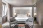 A modern hotel room with a large bed, two bedside tables with lamps, two gray armchairs, a small table with books, and a large mirrored wardrobe, decorated in neutral tones with soft lighting.
