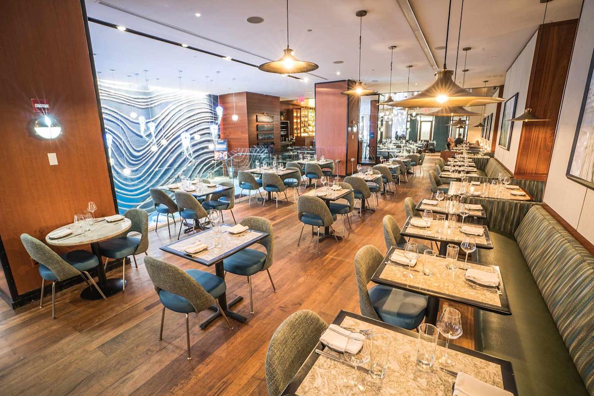 Modern restaurant interior with wooden floors, marble tables, green upholstered chairs, and banquette seating. Stylish lighting hangs from the ceiling, and a wavy blue accent wall decorates the back of the room. Tables are set for dining.