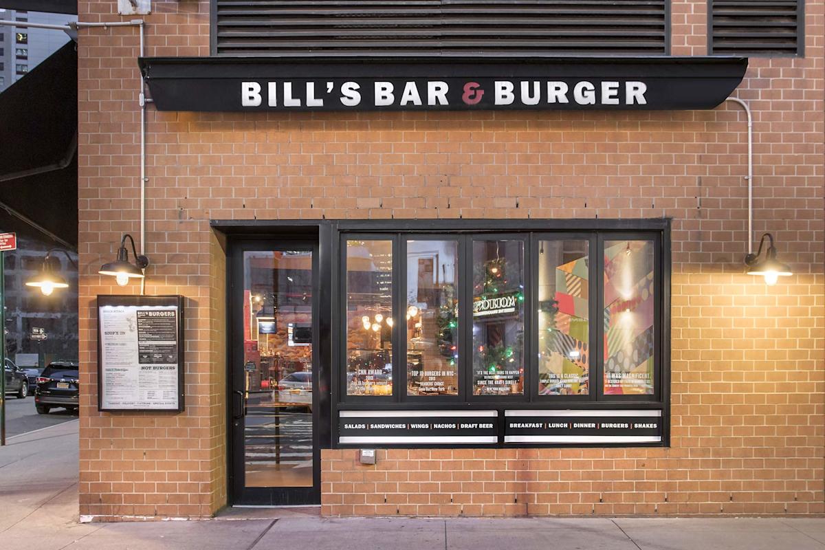 Exterior of Bill’s Bar & Burger, showing a brick building with black awning and white lettering, large windows revealing a lit interior, two wall-mounted lamps, and a menu posted outside.