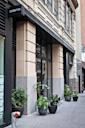 Street view of the entrance to a restaurant named Cosme, featuring large glass doors and several black planters with green plants arranged along the sidewalk. The building has beige stonework and tall windows.
