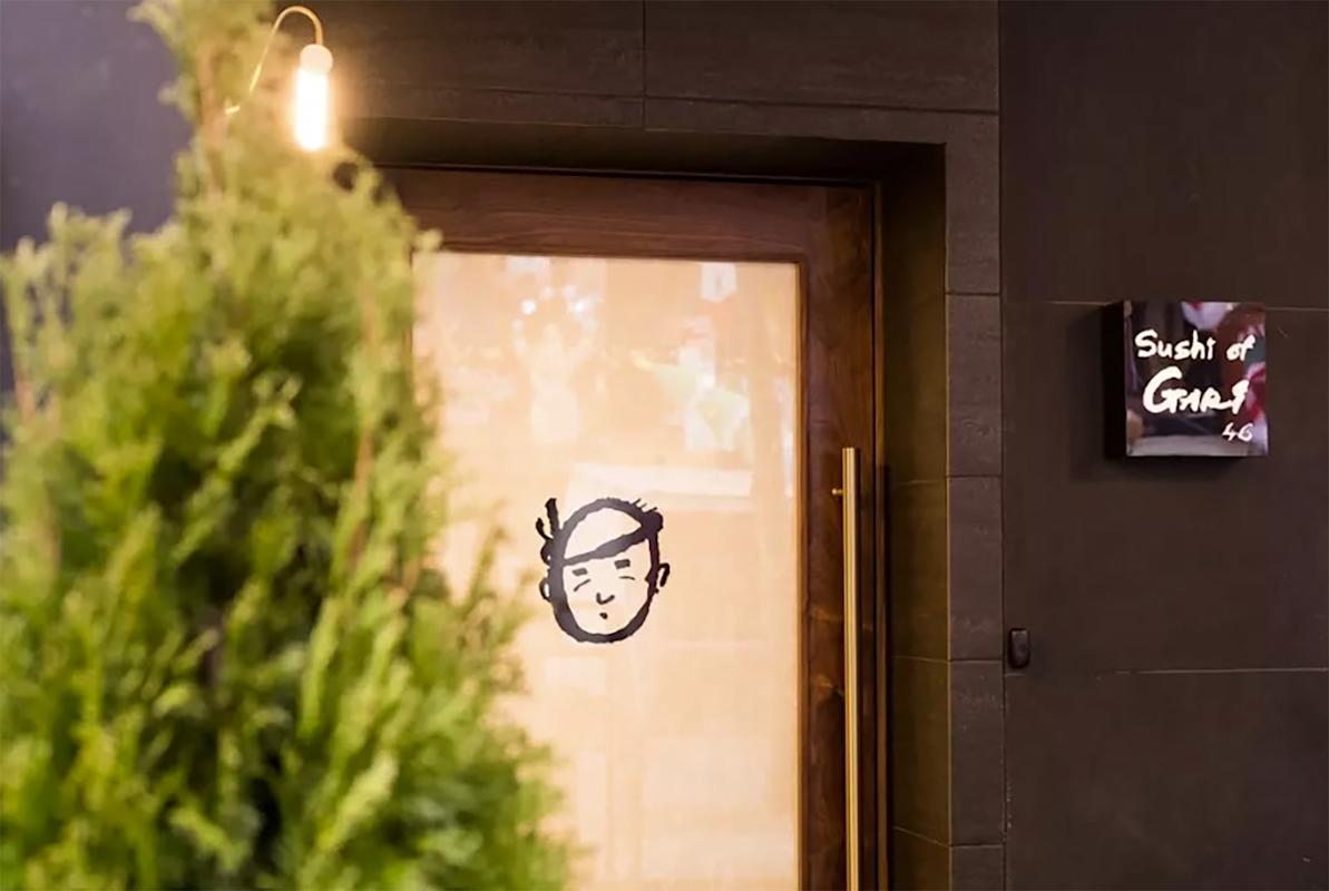A blurred green plant in the foreground partially obscures a wooden door with a cartoon face on frosted glass. A small sign next to the door reads "Sushi at Gari.