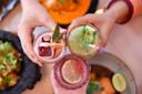 Three colorful beverages held up in a toast. Each glass has garnishes: a lime wedge, herbs, and salted rims. The background features dishes with vibrant food, including vegetables and lime. Hands are visible, with one wearing a purple sweater.