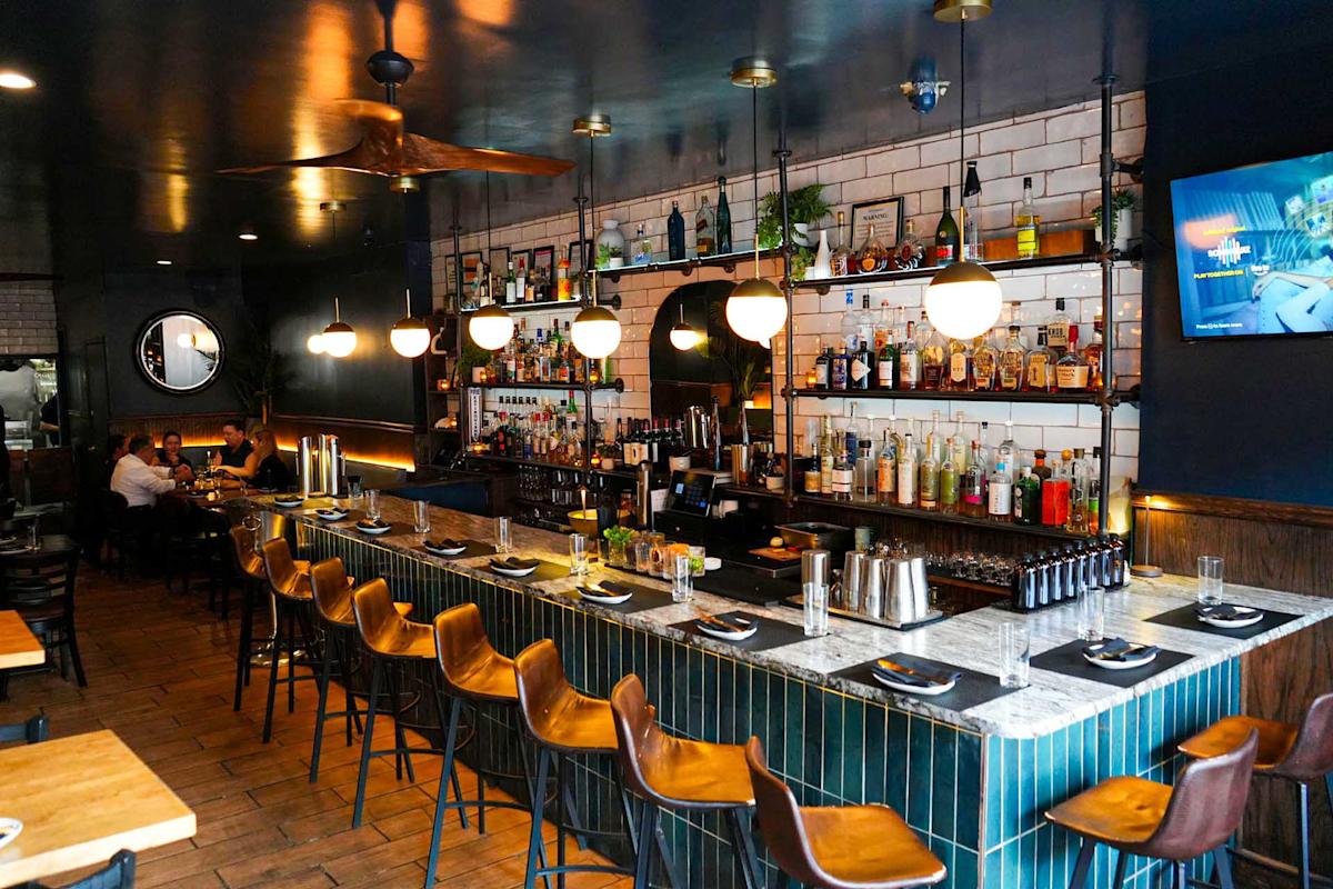 A modern bar with a marble countertop, brown barstools, and shelves of liquor bottles. Warm lighting and a TV on the wall create a cozy atmosphere; a few people are seated in the background.