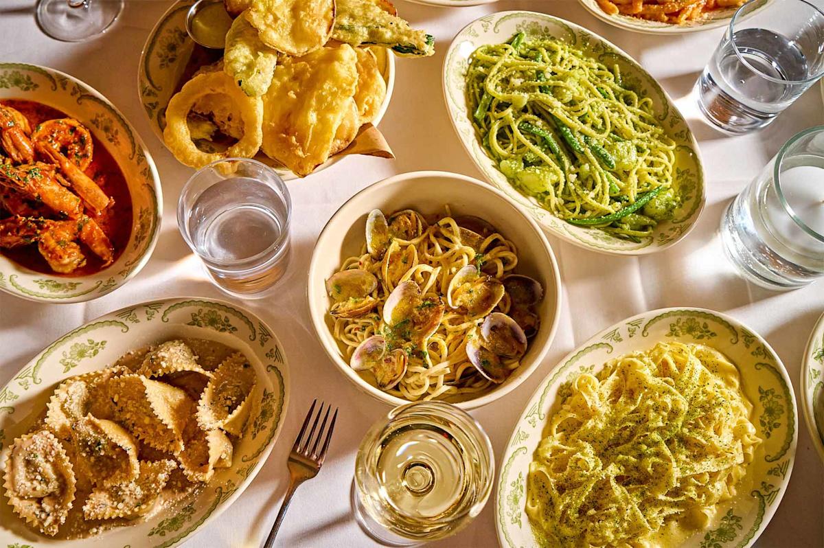 A table set with various Italian pasta dishes, fried vegetables, shrimp in tomato sauce, ravioli, and glasses of water and white wine, all served on patterned plates and bowls.