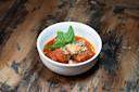 A white bowl filled with meatballs in tomato sauce, garnished with grated cheese and a sprig of fresh basil, sits on a rustic wooden table.