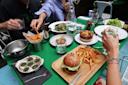 A group of people share a meal at a green table with burgers, fries, escargot, salad, pie, and drinks. Hands reach for food, and a small dog sits on one person's lap. Plates, glasses, and utensils are scattered around.