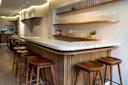 Modern bar interior featuring a terrazzo countertop with wooden stools arranged neatly around it. Shelves with an assortment of glassware are mounted on the wall. Warm lighting enhances the cozy ambiance.