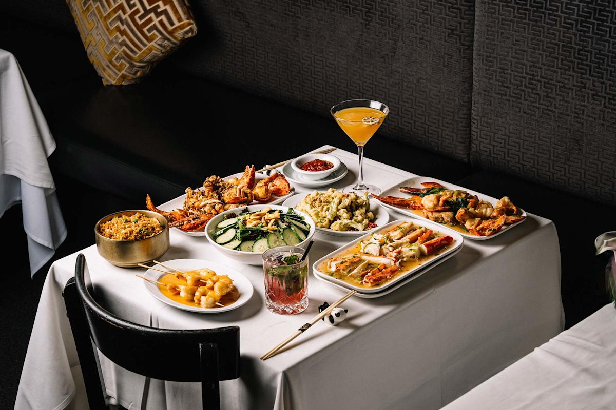 A white-clothed table set with various seafood dishes, vegetables, rice, and two cocktails, in a dimly lit restaurant with black seating and patterned pillows.