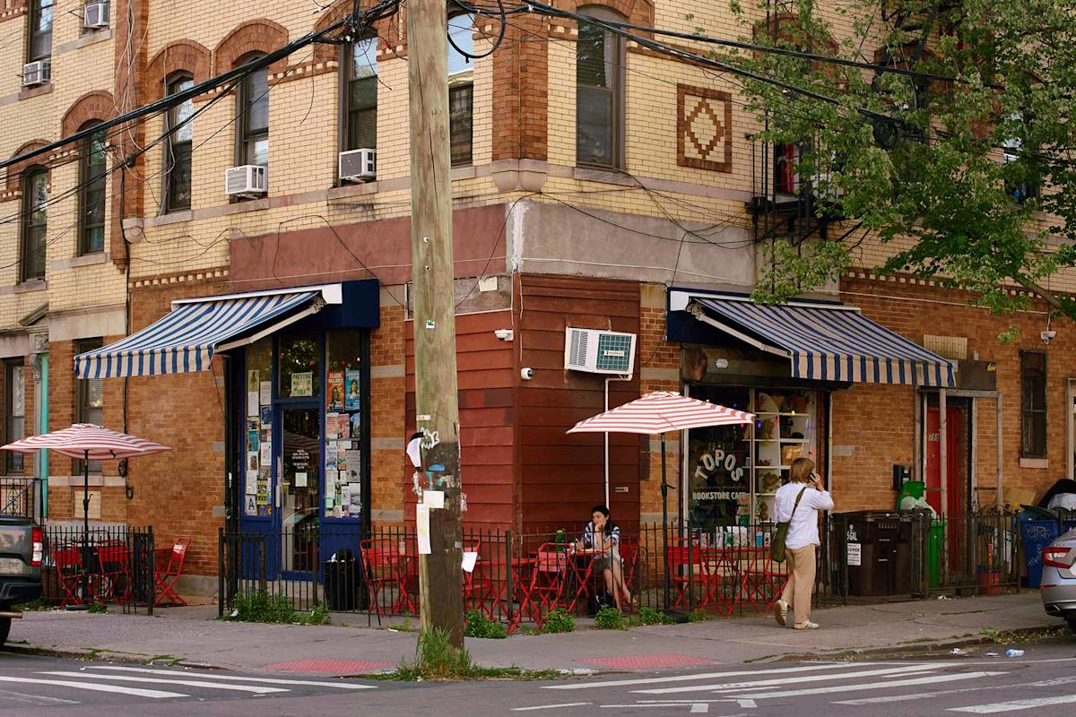 A street corner cafe with red chairs and tables outside, striped umbrellas, people sitting and walking, and brick buildings in the background.
