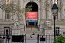 The image shows the entrance of the National Museum of the American Indian. It features grand steps, ornate statues on either side, and a large red banner above the entrance. Leafy trees and a classic lamppost frame the scene.