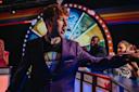 A man in a purple blazer excitedly gestures while standing in front of a colorful spinning game show wheel, with cheering people behind game podiums in a lively studio setting.