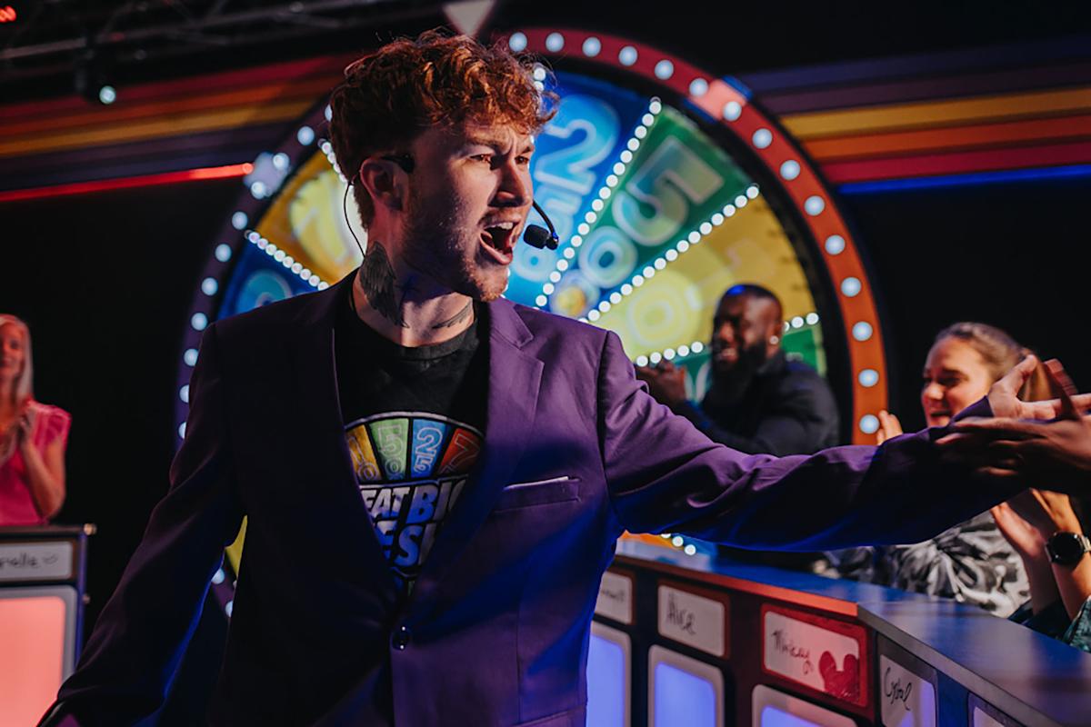 A man in a purple blazer excitedly gestures while standing in front of a colorful spinning game show wheel, with cheering people behind game podiums in a lively studio setting.