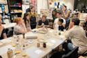 A group of people of diverse ages sit around a table covered with art supplies in a well-lit room, while one person demonstrates a technique; others observe attentively, some standing and some seated.