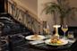 Two elegantly plated dishes on a green marble table, accompanied by wine glasses and a small lamp. A staircase with "New York" on the railing is in the background, alongside a potted plant.