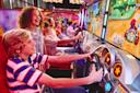 A group of kids happily play a racing video game at an arcade, focusing on the screen and holding steering wheels, with colorful lights and game graphics in the background.