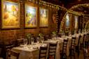 A warmly lit restaurant with white tablecloths, wooden chairs, wine glasses, and green-shaded lamps. Colorful landscape paintings hang on a brick wall adorned with string lights.