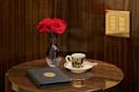 A small round wooden table holds a vase with two red roses, a black and gold notebook, and an ornate cup and saucer with a spoon. The background features glossy dark wood and a panel with gold switches.