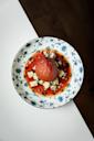 A round dessert, possibly poached fruit, sits on a floral-patterned plate with red syrup and crumbled cheese, photographed from above on a white and dark brown surface.