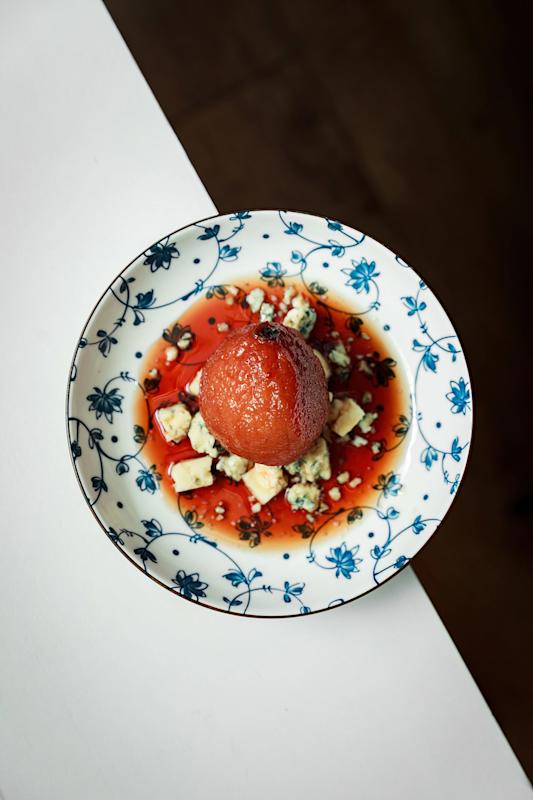 A round dessert, possibly poached fruit, sits on a floral-patterned plate with red syrup and crumbled cheese, photographed from above on a white and dark brown surface.