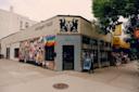 Corner view of Sanger Hall, a bar with black lion silhouettes and a heart sign on the exterior, a wall covered in posters and a rainbow flag, and a chalkboard sign by the entrance; a 99-cent store is nearby.