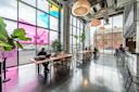 Modern cafe with high ceilings, large windows with colorful tinted panels, hanging lights, and potted plants. Several people sit at wooden tables, working on laptops or talking. An industrial brick building is visible outside.