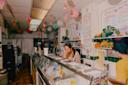 A woman stands behind the counter of a brightly decorated ice cream shop with colorful hanging paper lanterns and menu boards on the walls. Another person is behind the counter, partially turned away.
