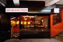 Street-level view of the entrance to a restaurant called "See No Evil Pizza," with a lit sign above glass doors. Warm lighting and tables are visible inside through large windows.