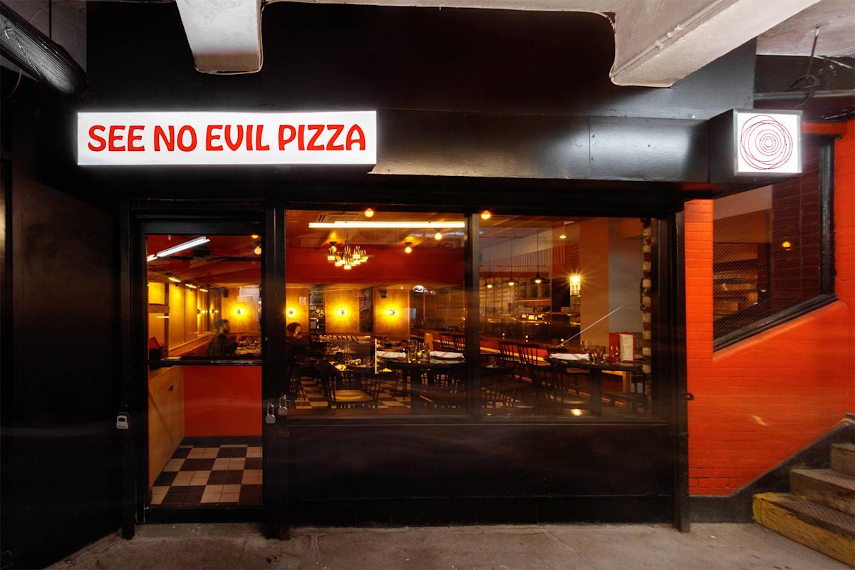 Street-level view of the entrance to a restaurant called "See No Evil Pizza," with a lit sign above glass doors. Warm lighting and tables are visible inside through large windows.