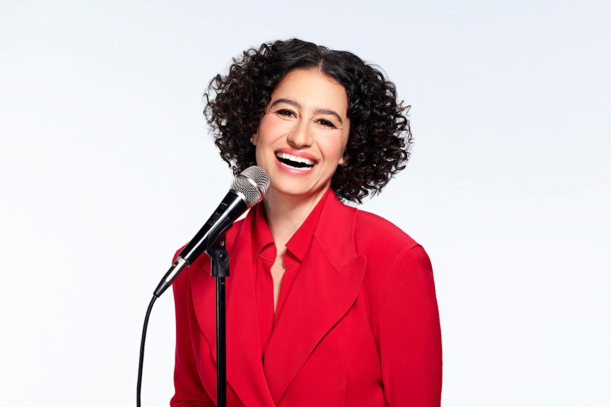 A woman with long braids and manicured nails poses confidently with arms crossed over a vintage microphone, set against a solid mustard yellow background.