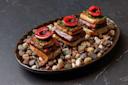 Three gourmet appetizers are arranged on a dark oval plate filled with small pebbles. Each appetizer features a piece of meat, topped with herbs and a cherry half, all resting on a small bread square.
