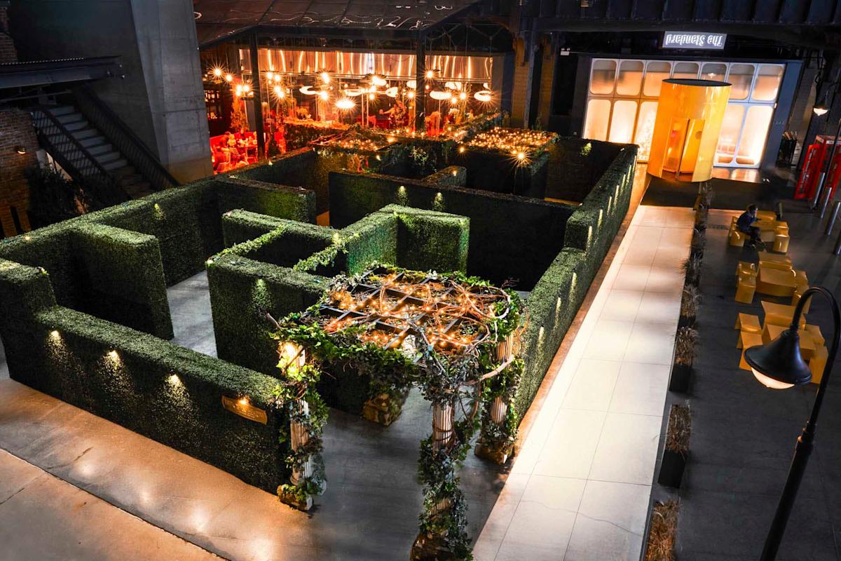 An indoor hedge maze with lush green walls is lit by warm overhead lights. There are tables and chairs set up along a pathway beside the maze. A glass entrance and seating area are visible in the background.