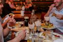 People are sitting at a restaurant table, enjoying a meal and drinks. Plates of food and glasses, including a pint of dark beer, are visible. One person holds a slice of pizza, and another is laughing while holding a sandwich. The atmosphere is lively.