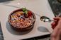 A person holding a spoon approaches a dish of crème brûlée on a white plate. The dessert is garnished with powdered sugar, fresh raspberries, and a mint leaf, set on a dark textured surface.