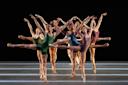 Eight ballet dancers in colorful leotards perform on stage, extending one leg and both arms gracefully in unison, creating a symmetrical and dynamic formation against a dark background.
