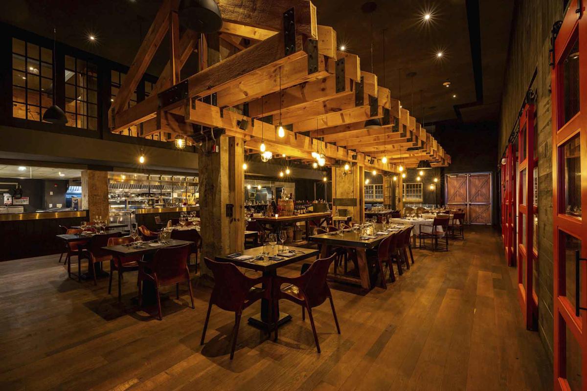 A warmly lit restaurant interior features wooden beams, exposed ceilings, large windows, and neatly set tables with red chairs on a hardwood floor. The open kitchen is visible in the background.