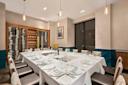 Elegant dining room with a long, white-clothed table set for a meal, surrounded by cushioned chairs. Wine racks line the back wall, and modern pendant lights hang from the ceiling.