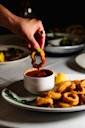 A hand dips a piece of fried calamari into a small bowl of red marinara sauce on a white plate, with more calamari and a lemon wedge on the side.