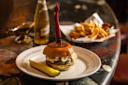 A cheeseburger with a red-handled knife through the bun is served on a plate with a pickle. In the background, there are French fries in a basket, a bottle of beer, and sauce on a marble table.