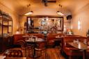 Cozy restaurant interior with brown leather booths, wooden tables set for dining, warm lighting, large mirror on the wall, and a vase of white flowers as a centerpiece.