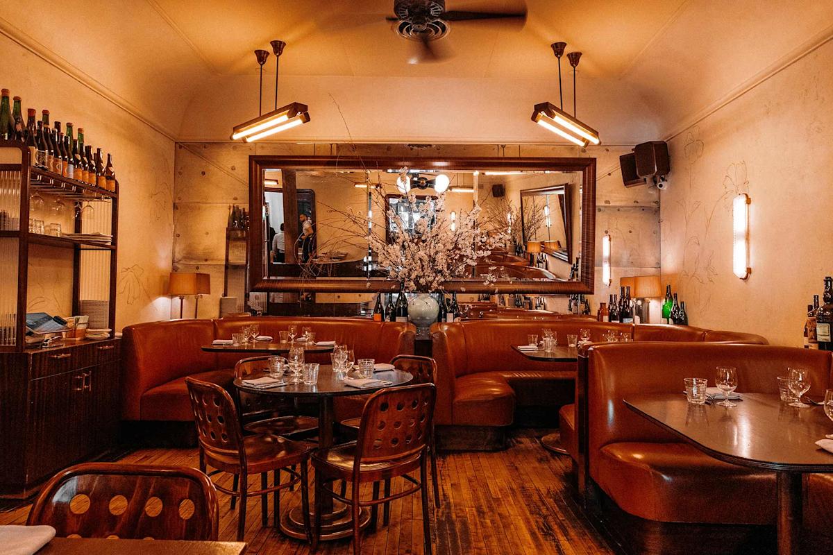 Cozy restaurant interior with brown leather booths, wooden tables set for dining, warm lighting, large mirror on the wall, and a vase of white flowers as a centerpiece.
