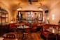 Cozy restaurant interior with brown leather booths, wooden tables set for dining, warm lighting, large mirror on the wall, and a vase of white flowers as a centerpiece.