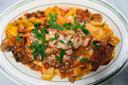 A plate of pappardelle pasta topped with a rich meat and tomato sauce, garnished with chopped parsley and grated cheese, served on an oval white dish with a green rim.